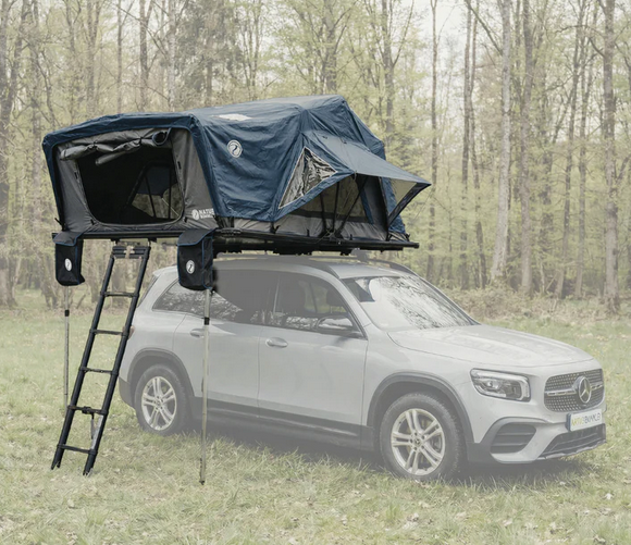 Ein silberner Mercedes SUV mit einem dunkelblauen Dachzelt und aufgeklappter Leiter steht auf einer Wiese vor lichten grünen Bäumen